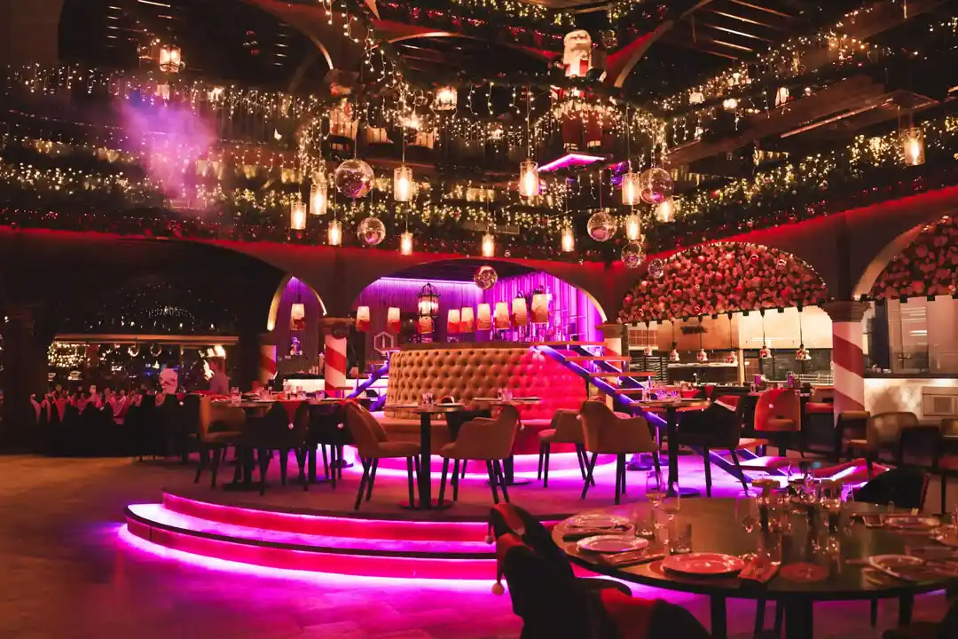 A lavishly decorated restaurant interior with festive Christmas lights, garlands, and vibrant magenta floor lighting.