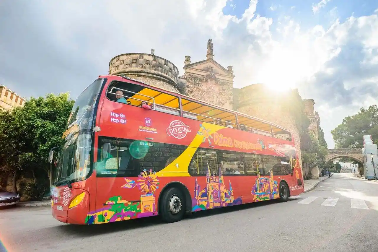 A red double-decker tourist bus drives on a road near a historic stone castle surrounded by green trees under a blue sky.