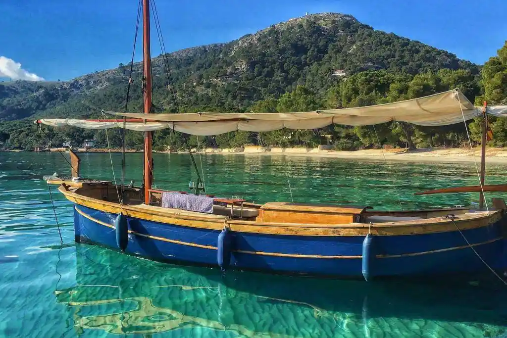 Traditional blue wooden boat floating on turquoise water near a beach and a forested hill.