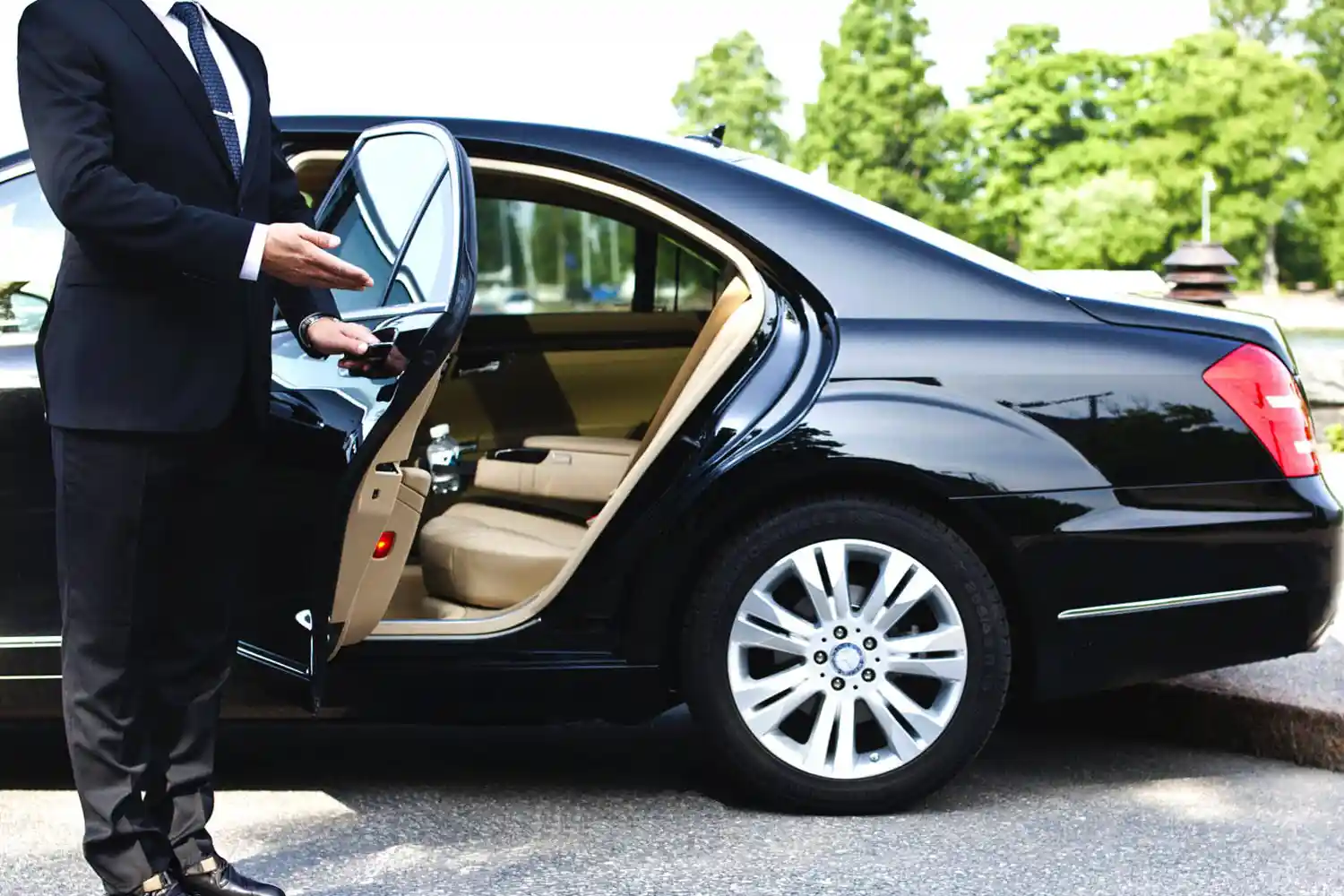A chauffeur in a suit opens the rear door of a luxury black sedan car on a sunny day.
