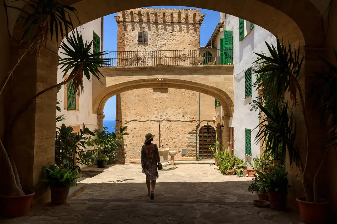 A person walks through a historic stone courtyard with arches and a medieval tower in the background.
