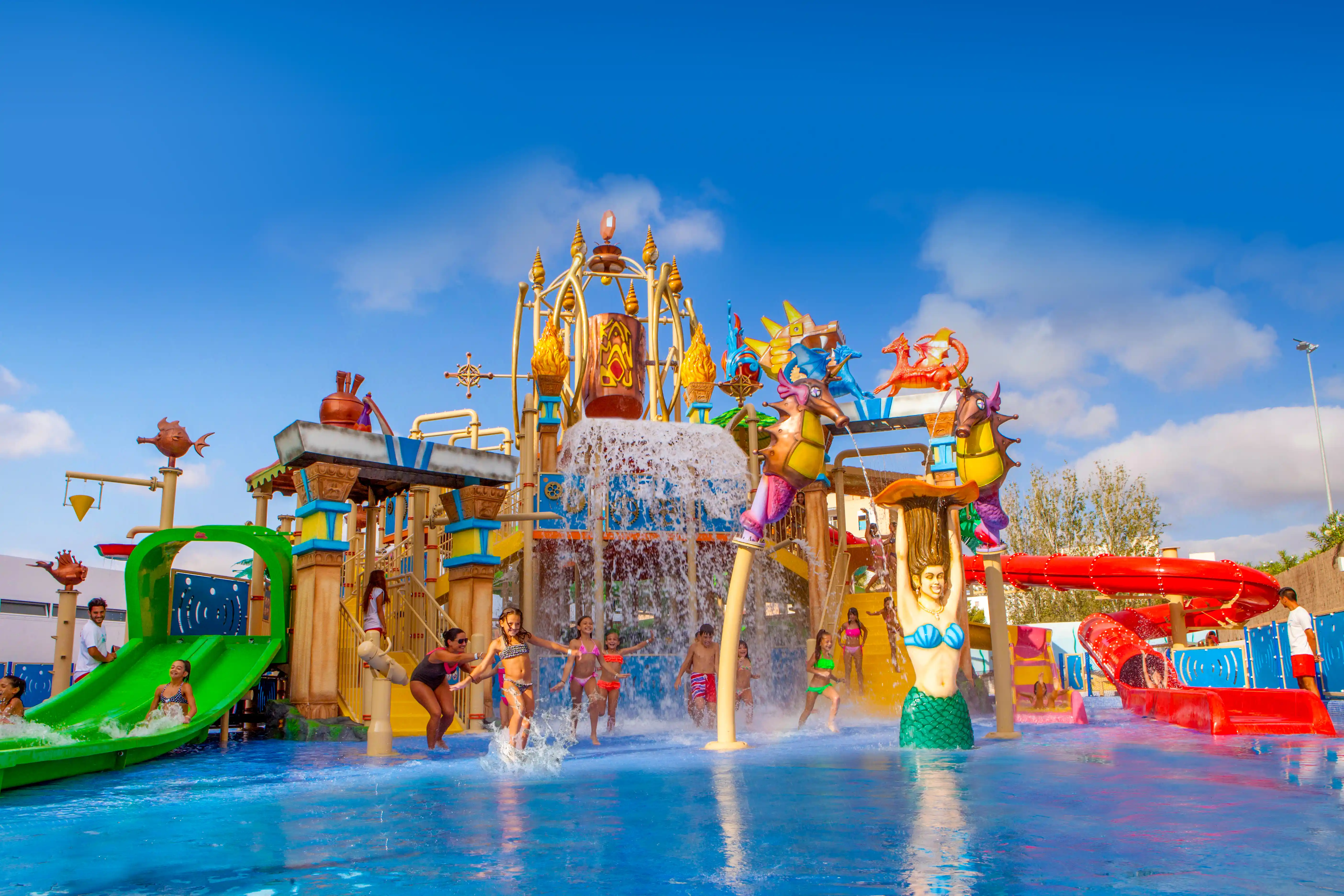Children play in a colorful water park with slides, a large bucket splash, and mythical decorations under a blue sky.