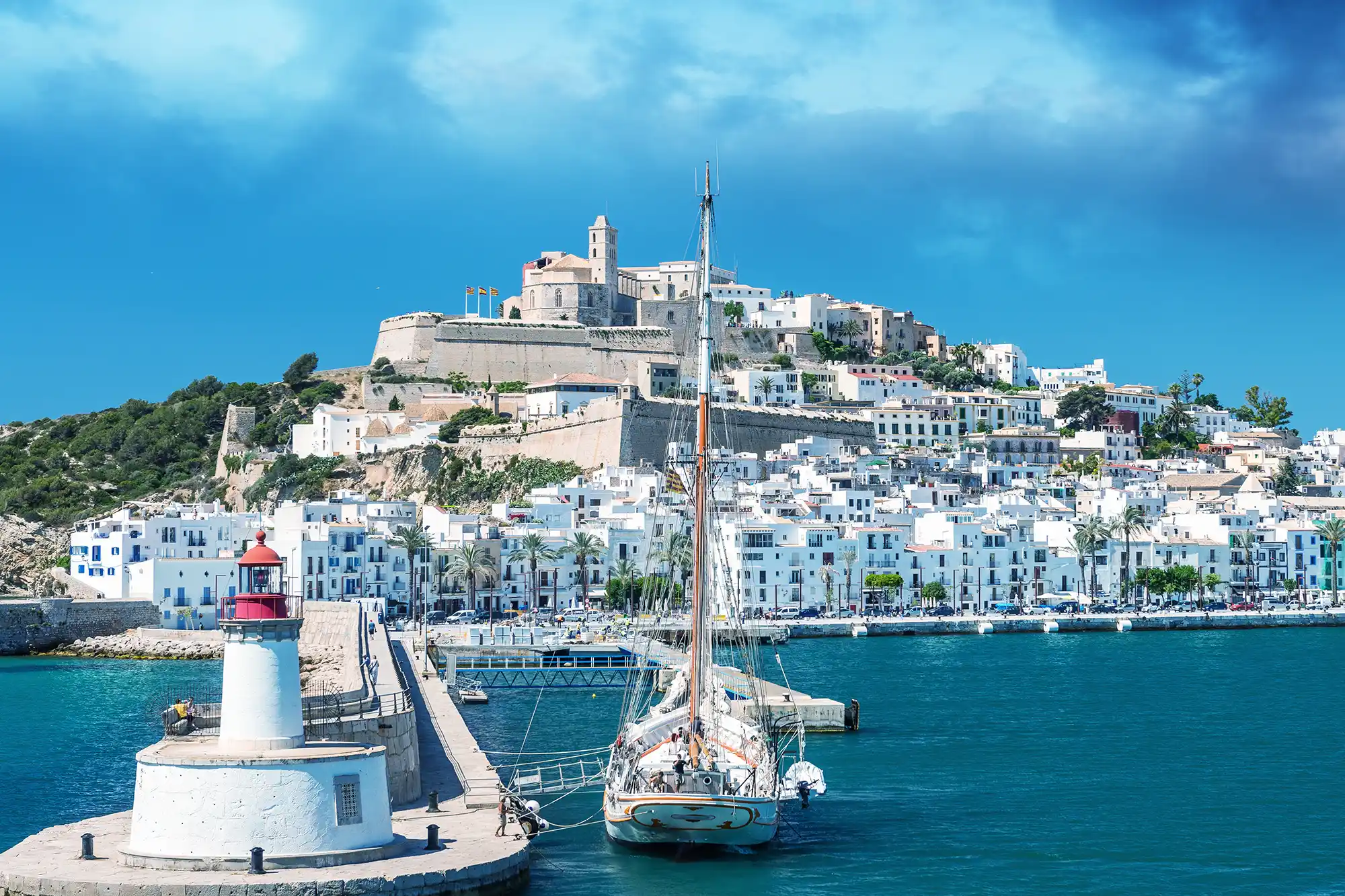 Scenic view of Ibiza old town with a lighthouse, anchored boat, and the historic fortress on the hill.